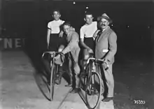 Photographie en noir et blanc montrant deux cyclistes montés sur leurs vélos, à l'arrêt sur une piste, s'appuyant sur deux hommes en civil.