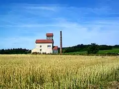 Un bâtiment industriel avec une haute cheminée, isolé au beau milieu de champs.