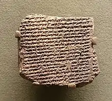 Tablette d'argile beige de forme relativement carrée couverte de caractères cunéiformes attachée à un mur à l'aide de quatre supports.
