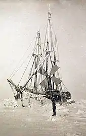  Vue de face légèrement décalée d'un navire couvert de givre, entouré de monceaux de glace. Un personnage solitaire se trouve debout sur la glace à côté du navire.