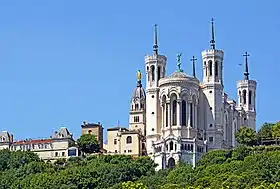 Photographie couleur montrant la basilique, au sommet de la colline, avec ses quatre tours et la statue de la Vierge, prise depuis le centre-ville de Lyon