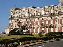 Vue d'un grand immeuble à trois étages ; façades couleur brique ou pierre et brique ; toiture en ardoises.