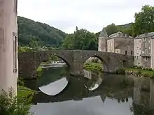 Photo couleur d'un pont à deux arches se reflétant dans une rivière. Il est situé dans un village verdoyant.