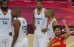 Quatre joueurs français en blanc, les mains sur les hanches. Un joueur espagnol lève le poing en signe de victoire.