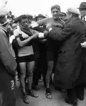 Photographie en noir et blanc montrant deux cyclistes enfilant un maillot à l'arrivée d'une course, entourés par la foule.