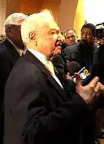 Au premier plan, Frank Gehry de profil est interviewé. Au second plan, caméra et autres journalistes.