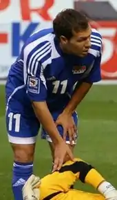 Un joueur de football au maillot bleu est debout et se penche vers le sol. Vu de face, il regarde à sa gauche.