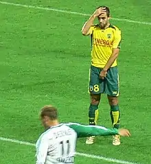 Photo de deux joueurs de football. L'un est flouté et s'apprête à tirer. L'autre est en jaune et se protège du tir.