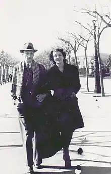 Un couple, bras dessus, bras dessous, marche face à l'objectif. L'homme porte costume, chapeau et écharpe. La femme est tout sourire