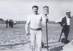 Portrait d'un homme à moustache portant une tunique claire et tenant en main une barre surmontée de poids
