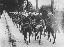 Photo en noir et blanc d'une colonne de cavaliers, par rang de trois, portant casque à crinière et la lance à l'étrier.