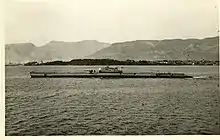 Photographie en sepia d'un sous-marin naviguant à proximité d'un rivage montagneux.