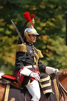 Buste d’un homme à cheval en uniforme noir, rouge et or, le sabre à la main, et coiffé d’un casque doré orné de plumeaux rouges et d’une longue que de cheval noire.
