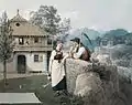 Filles avec arrosoir et paysan avec la faux devant une maison.