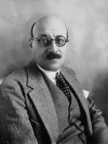 Portrait en buste d'un homme, corps de trois-quarts et tête de face, chauve avec une moustache et portant des lunettes cerclées.