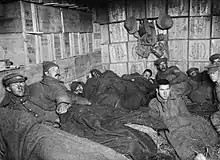 Photographie de l'intérieur d'un abri avec des murs composés de caisses de bois. Une dizaine d'hommes sont allongés avec des couvertures sur un sol couvert de paille