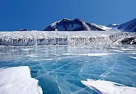 Vue depuis un lac gelé couvert de glace bleue et bordé par un glacier descendant en pente faible d'une montagne.