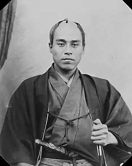 Photo noir et blanc. Homme au crâne dégarni, avec habit traditionnel japonais sombre et sabre à la taille. Fond blanc.
