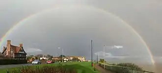 arc-en-ciel en demi-cercle sur ciel gris, une maison à gauche et la mer à droite séparées par une route et de la pelouse