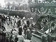 un char funèbre monumental est suivi par un cortège de personnages habillés en civil ou en militaires et avance au milieu de deux haies de spectateurs abrités par des parapluies et contenus par deux rangs de gendarmes