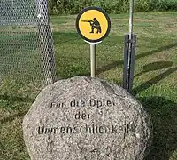 Boule de pierre portant inscription. « Für die Opfer der Unmenschlichkeit ». À l'arrière se trouve une section de clôture et un panneau jaune montrant un soldat agenouillé épaulant son arme.