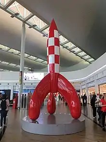 Fusée à damier rouge et blanc posé sur trois pieds, installée dans le hall d'un aéroport.