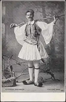 photographie noir et blanc : un homme moustachu en costume traditionnel (jupe) tenant un fusil