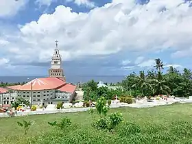 Photo en couleurs montrant l'arrière d'un bâtiment au toit rouge, surmonté d'un clocher blanc. En premier plan, de la pelouse et des cocotiers ; en arrière plan, la mer (océan Pacifique) et le ciel rempli de nuages blancs.