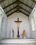 Photo d'une petite chapelle aux murs blancs, au toit en tôle avec une charpente en bois. à l'intérieur, une croix avec le Christ crucifié, et deux statues de chaque côté de personnages féminins