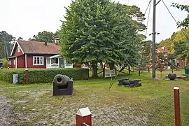 Petit village avec des maisons en bois peintes en rouge, entourées de verdure et un canon au premier plan.