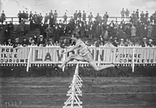 Photographie en noir et blanc d'un coureur à pied qui saute une haie dans un stade.