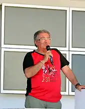 Photo en couleur d'un homme, debout, parlant devant un micro qu'il tient de la main droite.