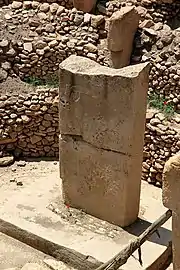 Deux pierres beiges rectangulaires posées l'une sur l'autre. Le dessus est visiblement cassé. À l'arrière-plan se trouve un ensemble de pierre superposées et enfoncées dans un mur.