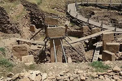 Vue en contre-plongé d'un pilier en T au milieu d'autre plus petites pierres levées. À l'arrière plan on voit une passerelle permettant de traverser le chantier de fouilles.