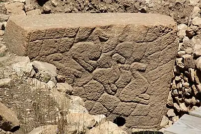 Pierre beige taillée rectangulaire, posée sur sa longueur et partiellement encastrée dans un muret fait de pierres. Sur la pierre il y a des animaux sculptés.