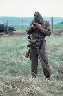 Garde est-allemand debout dans l'herbe et prenant une photo du photographe. Une clôture frontalière et un camion sont visibles derrière le soldat.