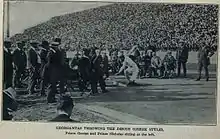 Photographie en noir et blanc montrant un athlète vêtu de blanc en train de lancer un disque devant des juges et de nombreux spectateurs.