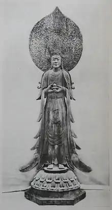 Vue de face d'une statue debout avec une grande robe et portant un petit récipient en face de son corps. Il y a une grande auréole derrière la tête de la statue.