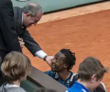Un dirigeant en costume-cravate au premier rang d'une tribune se penche sur un court de tennis pour féliciter un joueur.