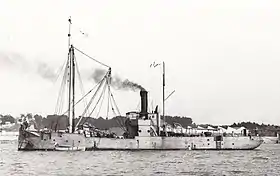 Photographie noir et blanc. Profil babord d’un navire de servitude au mouillage avec une chaudière sous pression.