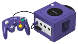 Console de jeux vidéo cubique de couleur violette, avec une manette filaire de même couleur équipée de boutons multicolores.