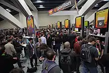Des joueurs font la queue devant différents stands de jeux vidéo oranges.