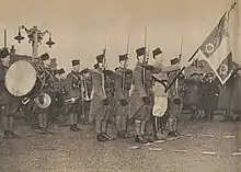 Militaires, en capote kaki et coiffés de la chéchia, présentant les armes autour de leur porte-drapeau. Derrière eux, des musiciens portant le même uniforme que la garde du drapeau.