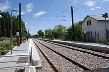 au centre les deux voies électrifiées, encadrées par deux quais avec le mobilier type de la ligne et à droite l'ancienne maison de garde barrière datant de l'origine de la ligne