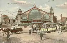 Carte postale colorisée représentant la façade de la gare d'arrivée et le carrefour avec des personnages, le kiosque et des tramways électriques.