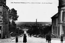 Carte postale ancienne de l'avenue de la gare à Noyal-sur-Vilaine avec au deuxième plan le bâtiment voyageurs de la gare de Noyal-Acigné et plus loin le bourg d'Acigné.