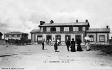 Carte postale 1900 en noir et blanc, avec au centre le bâtiment voyageurs vu de la place avec un groupe de personnage et à gauche des voitures à cheval en attente.