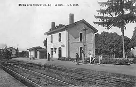 Carte postale photographie noir et blanc une locomotive à vapeur arrive en gare sur l'une des deux voies encadrées par deux quais, elle passe devant la halle à marchandises qui est à droite comme la lampisterie, les toilettes et le bâtiment voyageurs à deux entrées et un étage sous une toiture à deux pans en débords avec deux grandes cheminées en briques. Le chef de gare et deux autres personnages sont en attente sur le quai.