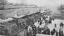 Photo noir et blanc prise depuis une passerelle, montrant un train de banlieue d'où descend une foule de voyageurs dont certains lisent des journaux.