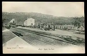 Carte postale noir et blanc. Une locomotive s'avance dans une gare de deux voies. Passagers sur le quai.
